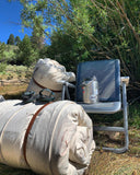 Two J bar D Cowboy Bedrolls laying in the grass next to a Yeti Chair and a Yeti Canteen. The background is a willow bush.